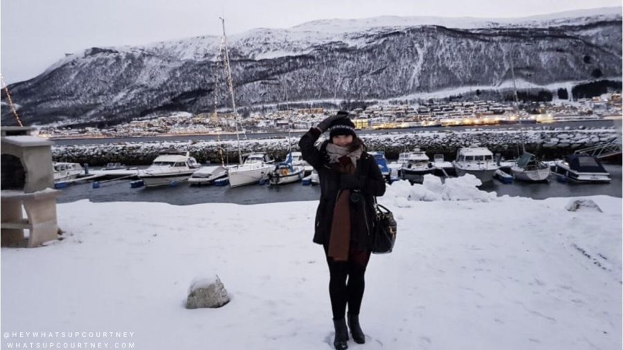Courtney (heywhatsupcourtney) from whatsupcourtney in front of a snowy mountain and boats in Tromso Norway