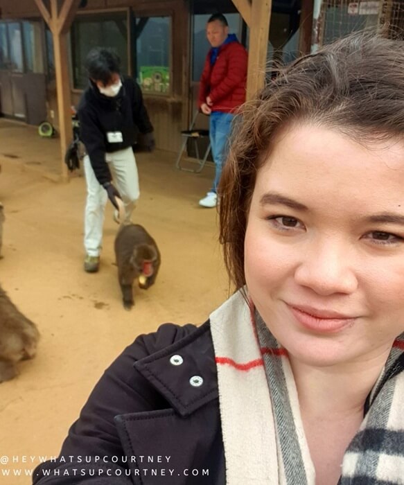 Courtney from whatsupcourtney.com posing in front of a Japanese Macaque monkey holding a treat and a keeper